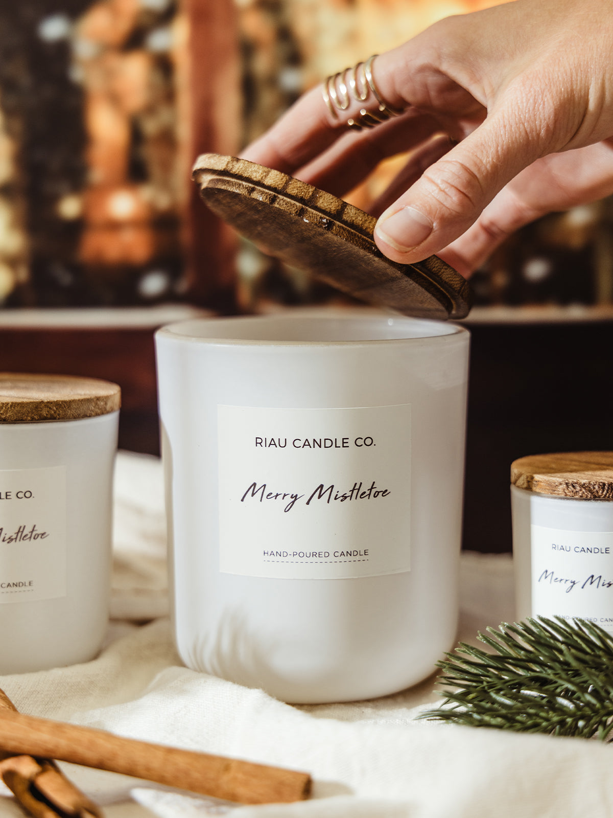 Close-up of a hand with gold rings placing a wooden lid on a large Merry Mistletoe candle by Riau Candle Co., styled with evergreens and cinnamon sticks.