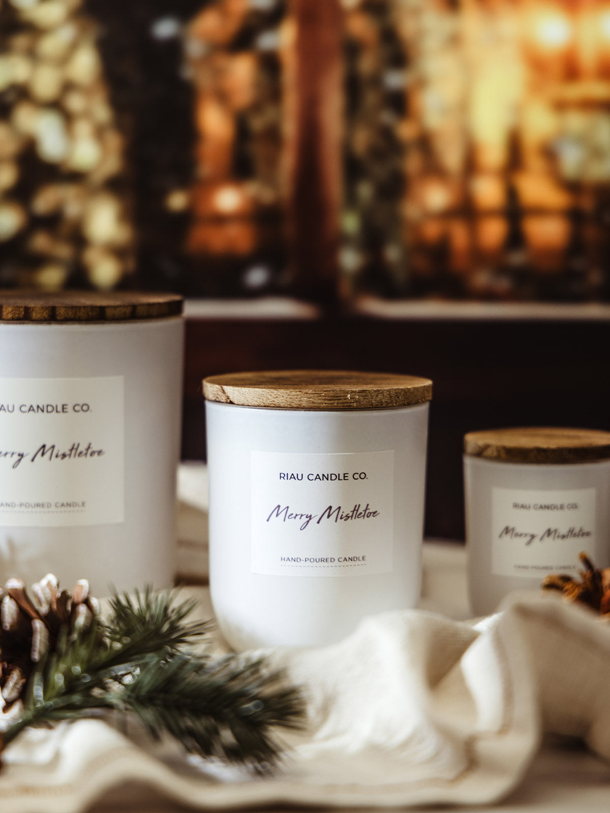 Medium Merry Mistletoe soy candle by Riau Candle Co. with wooden lid, styled among pinecones and greenery, glowing holiday lights softly blurred in the background.