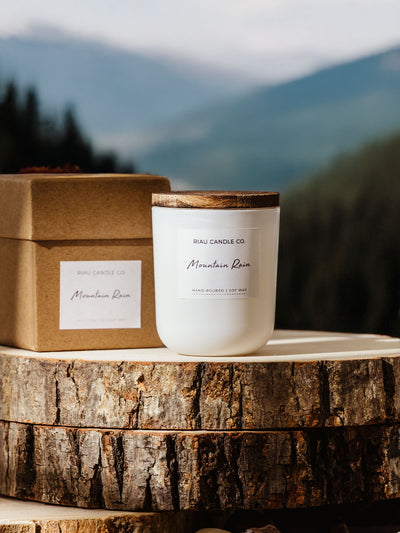 Riau Candle Co. Mountain Rain mini candle with a wooden lid, placed outdoors on natural wood slices with a matching kraft gift box.
