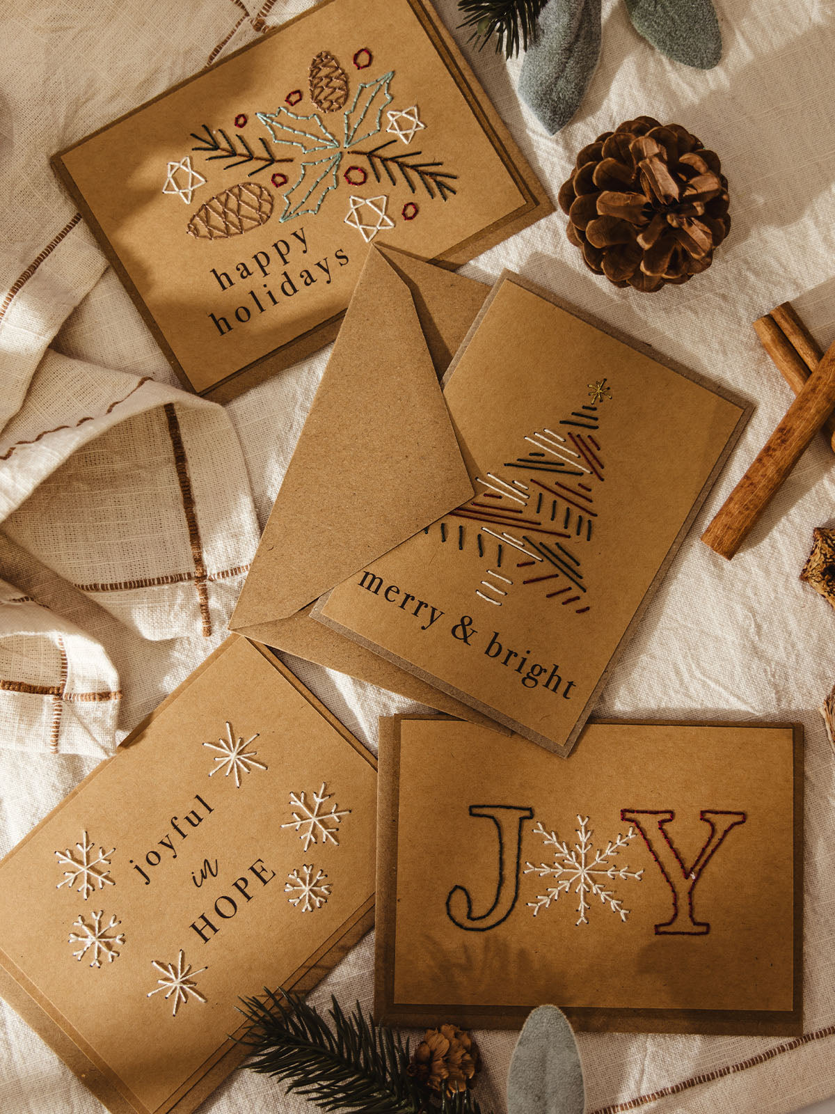 Flat lay of four brown kraft holiday cards with hand-stitched embroidery designs, surrounded by pinecones, cinnamon sticks, and evergreen sprigs on a cream cloth. The cards read: “happy holidays” with pinecones and holly leaves, “merry & bright” with a stitched Christmas tree, “joyful in HOPE” with snowflakes, and “JOY” with a snowflake as the O.