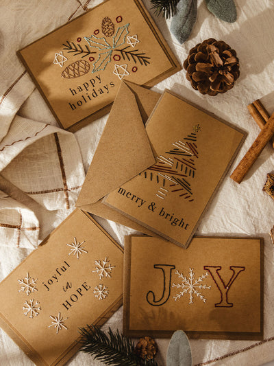 Flat lay of four brown kraft holiday cards with hand-stitched embroidery designs, surrounded by pinecones, cinnamon sticks, and evergreen sprigs on a cream cloth. The cards read: “happy holidays” with pinecones and holly leaves, “merry & bright” with a stitched Christmas tree, “joyful in HOPE” with snowflakes, and “JOY” with a snowflake as the O.