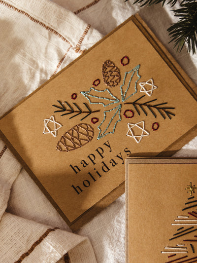 Brown kraft holiday card featuring embroidered pinecones, holly leaves, evergreen branches, and stars in colorful thread with the words “happy holidays” printed below.