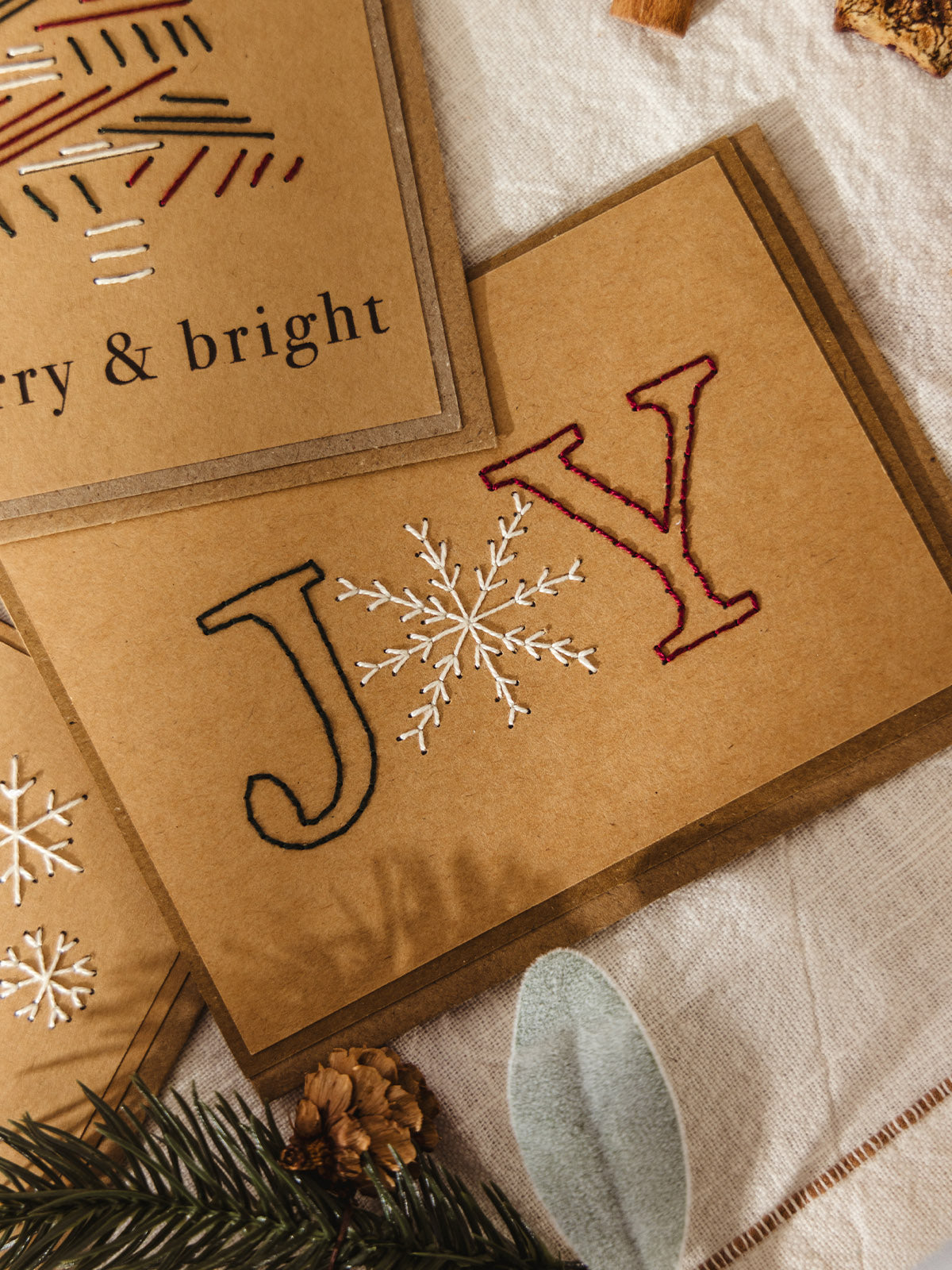 Brown kraft holiday card with stitched letters “J” and “Y” in green and red thread, and a white embroidered snowflake forming the “O” in “JOY.”