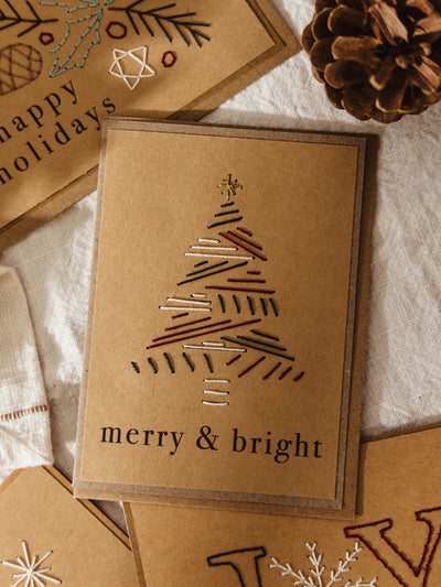Brown kraft holiday card with a minimalist embroidered Christmas tree made of colorful stitched lines, topped with a gold star, and the words “merry & bright” printed beneath.