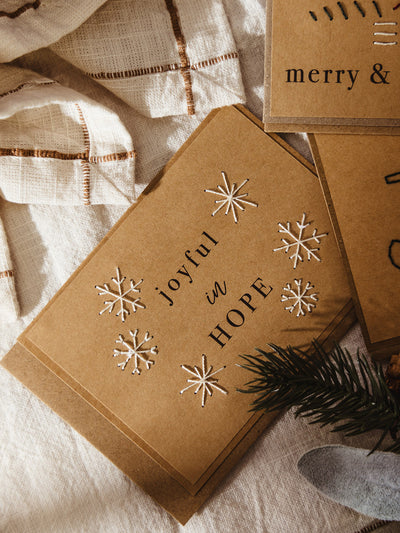 Brown kraft holiday card embroidered with white snowflakes and the phrase “joyful in HOPE” printed in black text across the center.