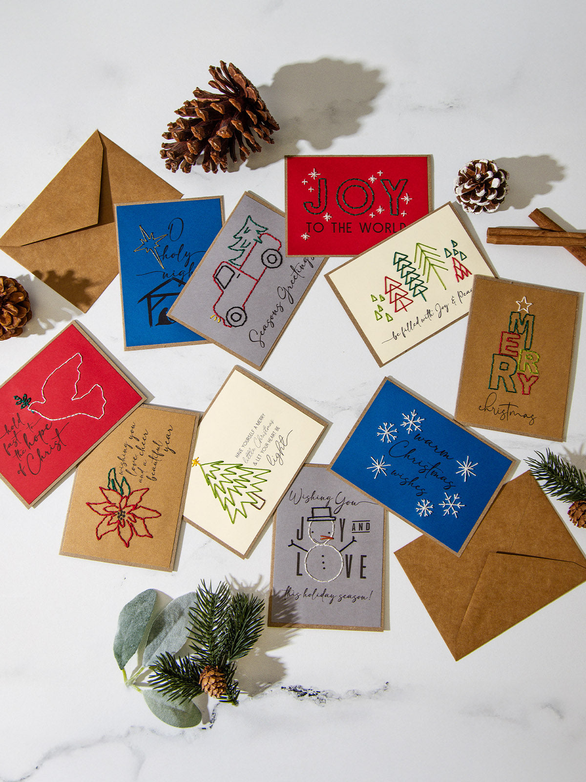 Flat lay of assorted handmade embroidered Christmas cards with brown kraft envelopes, pinecones, and greenery on a white marble surface. Designs include trees, snowflakes, a poinsettia, dove, nativity, and phrases like “Joy to the World” and “Season’s Greetings.”