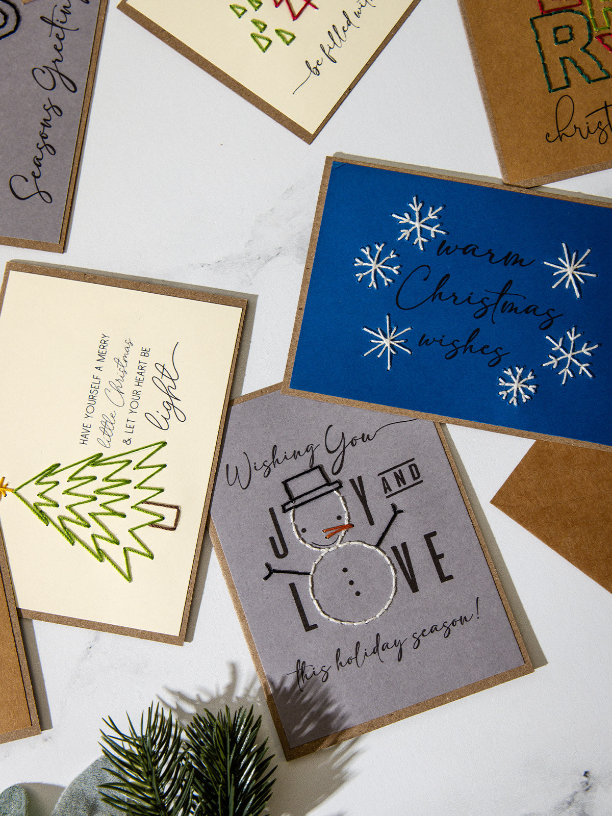 Close-up of embroidered Christmas cards featuring a stitched green tree with the phrase “Have yourself a merry little Christmas,” a gray snowman card reading “Wishing you joy and love,” and a blue card with white stitched snowflakes reading “Warm Christmas wishes.”