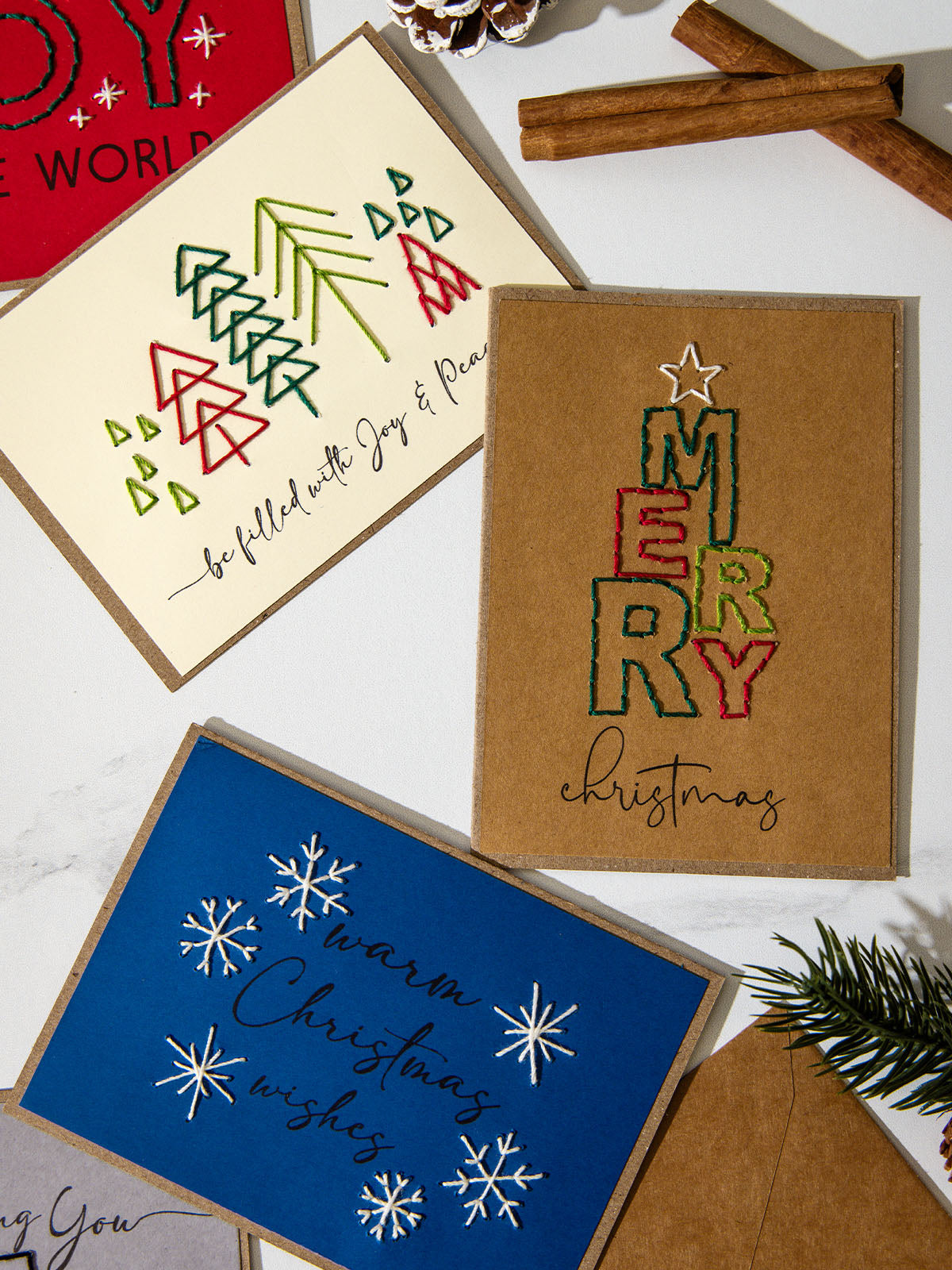 Embroidered holiday cards on a white background showing a modern tree pattern with “Be filled with joy and peace,” a kraft “Merry Christmas” stacked text design, and a blue card with stitched snowflakes reading “Warm Christmas wishes.”
