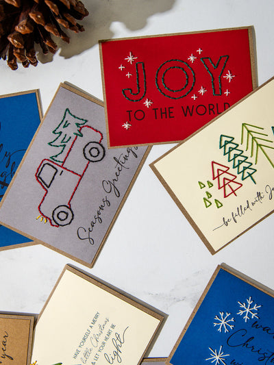 Overhead view of Christmas cards with hand-stitched designs including a red “Joy to the World” card, gray pickup truck carrying a tree with “Season’s Greetings,” and geometric embroidered trees with “Be filled with joy and peace.”
