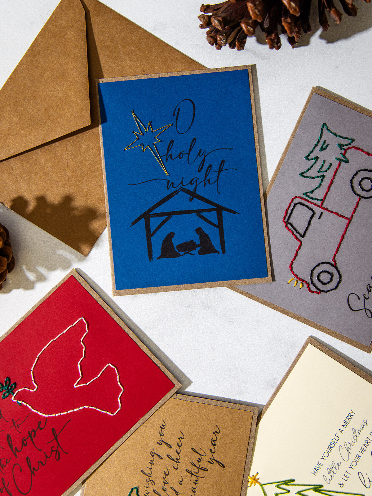 Close-up of blue “O Holy Night” Christmas card featuring a nativity silhouette with embroidered star, surrounded by other embroidered cards including a dove with “Hold fast to the hope of Christ” and a red truck carrying a Christmas tree.