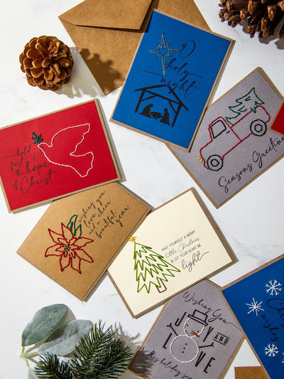Holiday card flat lay with embroidered designs including a red dove, poinsettia, green stitched Christmas tree, snowman, and nativity scene, styled with pinecones and greenery on white marble.