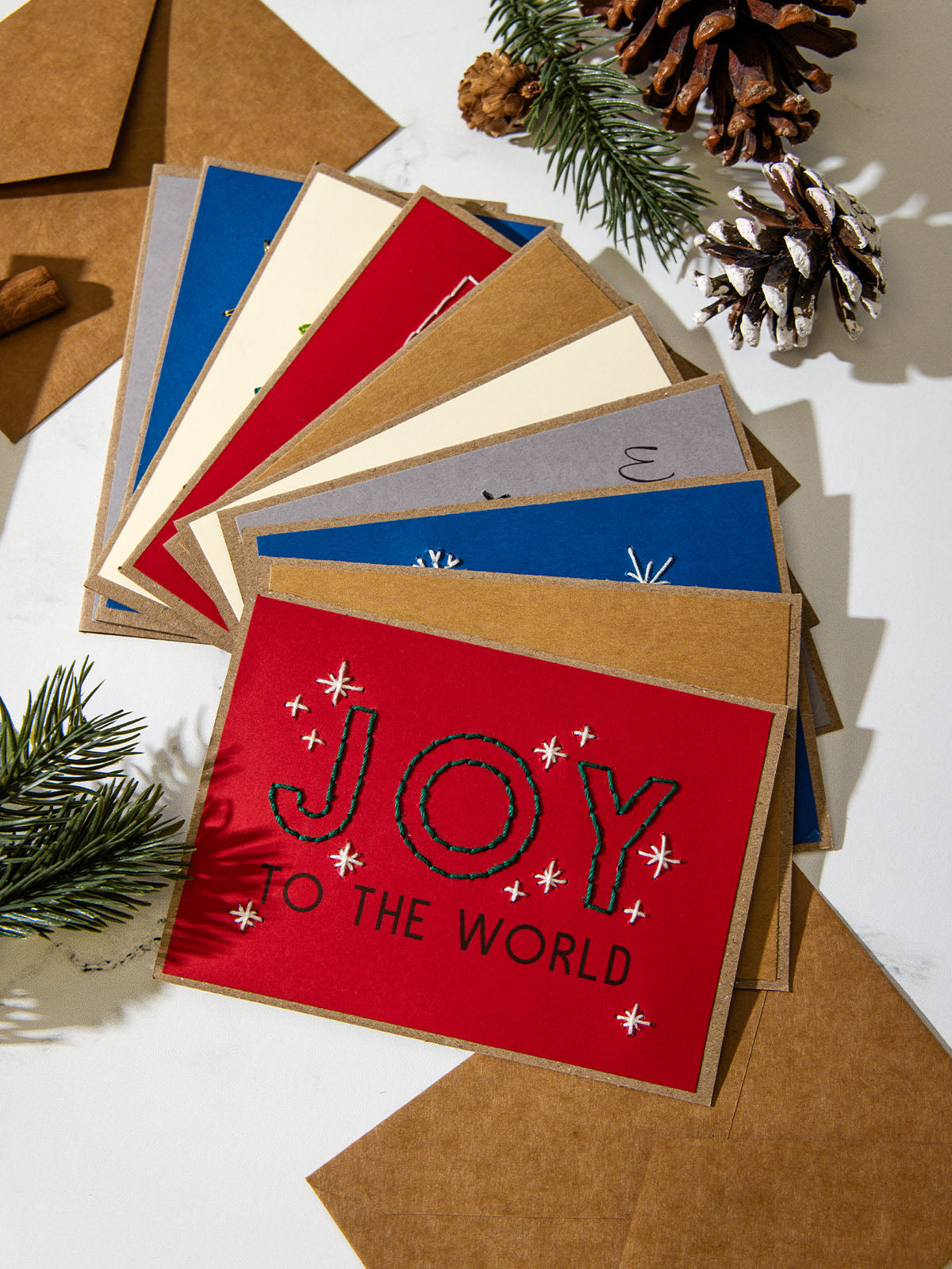 tacked set of colorful embroidered Christmas cards fanned out on white marble with pinecones and evergreen sprigs. Top card is bright red with green stitched text reading “Joy to the World.”