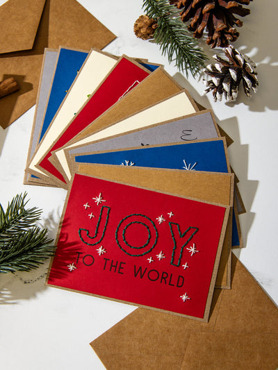 tacked set of colorful embroidered Christmas cards fanned out on white marble with pinecones and evergreen sprigs. Top card is bright red with green stitched text reading “Joy to the World.”
