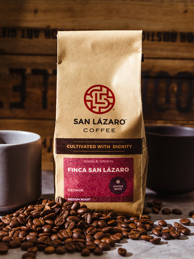 Bag of San Lázaro Coffee Finca San Lázaro whole bean medium roast, surrounded by roasted coffee beans and a ceramic mug, labeled “Cultivated with Dignity.”
