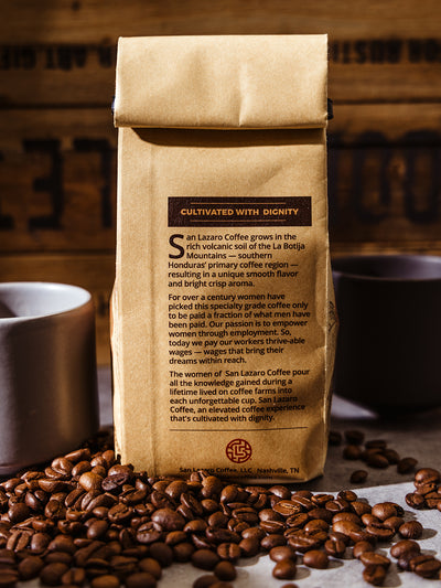 Back of San Lázaro Coffee bag detailing its origin from the La Botija Mountains in Honduras and the mission to empower women with fair wages, against a rustic wood backdrop.