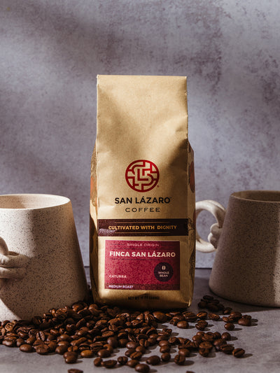 San Lázaro Coffee Finca San Lázaro bag styled between two speckled ceramic mugs on a concrete surface, with scattered coffee beans.