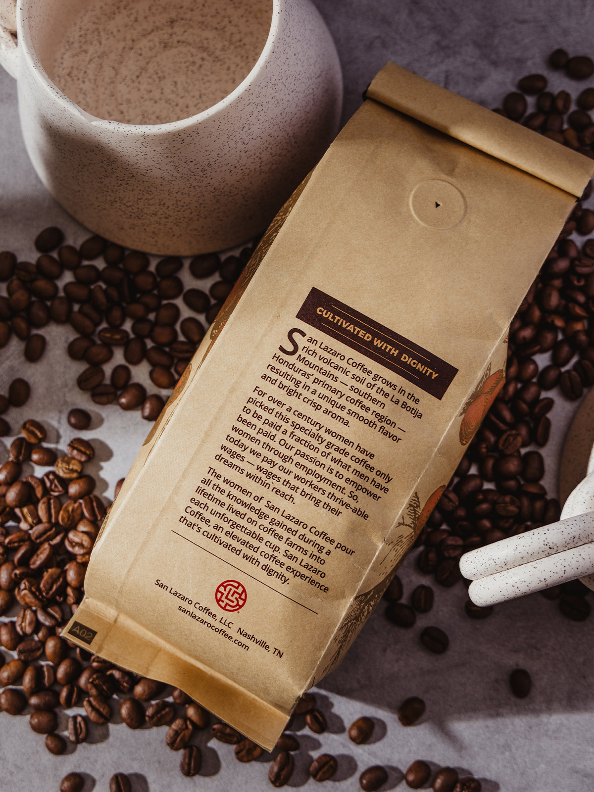 Detailed view of the back of San Lázaro Coffee bag lying flat among coffee beans and mugs, highlighting its story of ethical sourcing and women-led production.