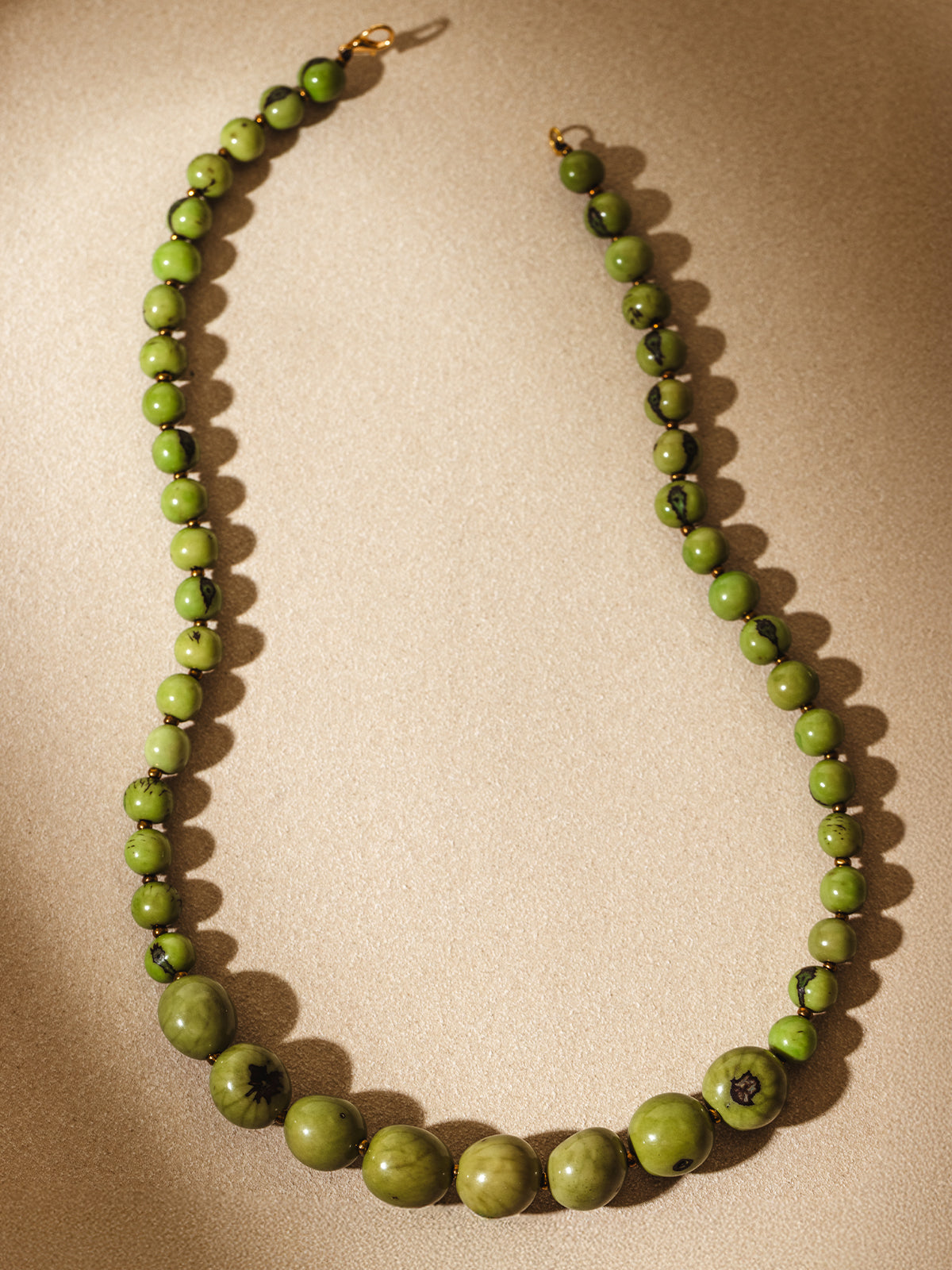 Flat lay of a handcrafted green beaded necklace with varying bead sizes and subtle marbling, arranged in a curve on a softly lit neutral background.