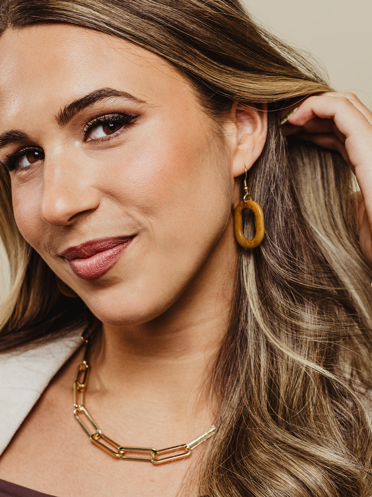 Close-up of a smiling woman wearing oval brown dangle earrings with a smooth finish, styled with voluminous hair and a gold chain necklace.