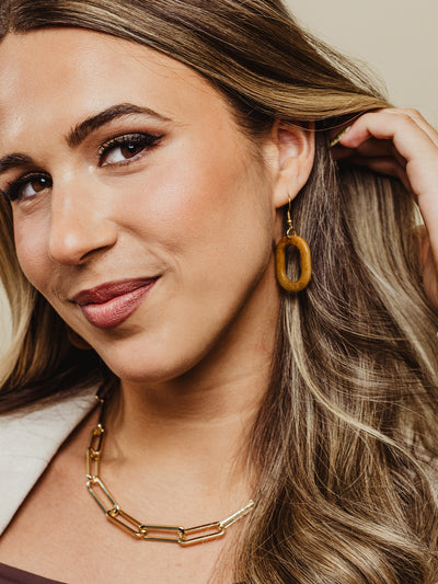 Close-up of a smiling woman wearing oval brown dangle earrings with a smooth finish, styled with voluminous hair and a gold chain necklace.