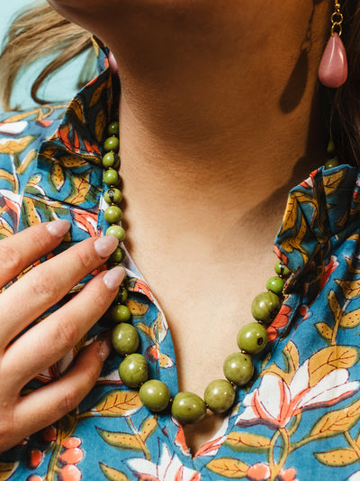 Close-up of a green beaded statement necklace with glossy round beads in various sizes, styled with a colorful floral blouse and pink teardrop earrings.