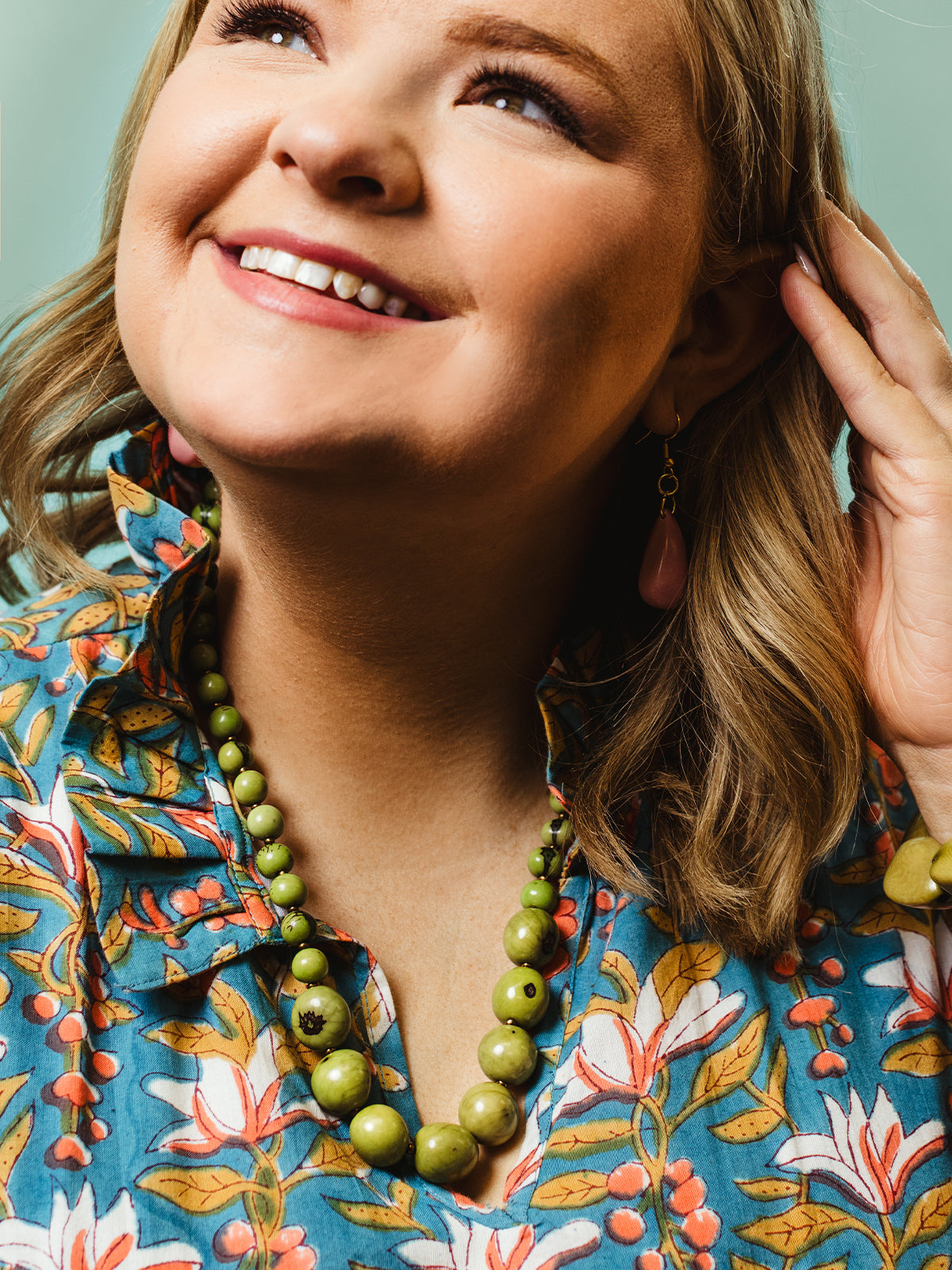 Portrait of a smiling woman wearing a bold green beaded necklace and matching bracelet, paired with pink teardrop earrings and a vibrant floral blouse.
