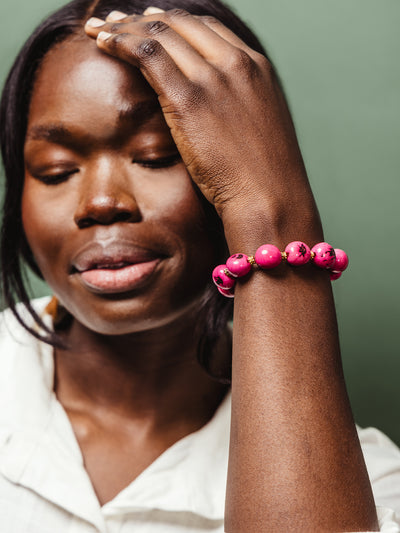 Portrait of a woman wearing a bold pink ceramic beaded bracelet with gold accents, her eyes closed and hand gently resting on her forehead, styled with a white blouse and a calm expression.