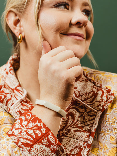 Smiling woman wearing a cream-colored bracelet and gold hoop earrings, dressed in a floral red and yellow patterned top, posing against a green background.