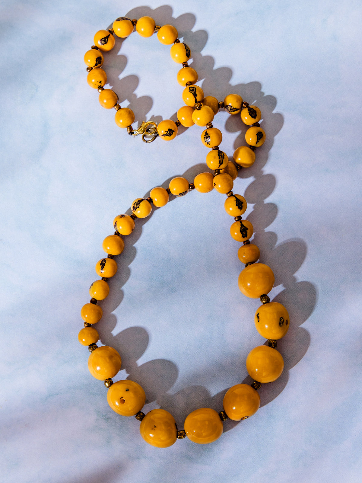 Vintage-style beaded necklace with round yellow beads and dark brown marbled details, displayed on a light blue background with shadows.