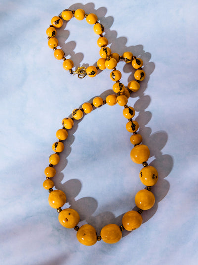 Vintage-style beaded necklace with round yellow beads and dark brown marbled details, displayed on a light blue background with shadows.