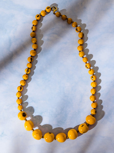 Handmade yellow beaded necklace with varying bead sizes and marbled accents, laid out in an oval shape on a soft blue surface.