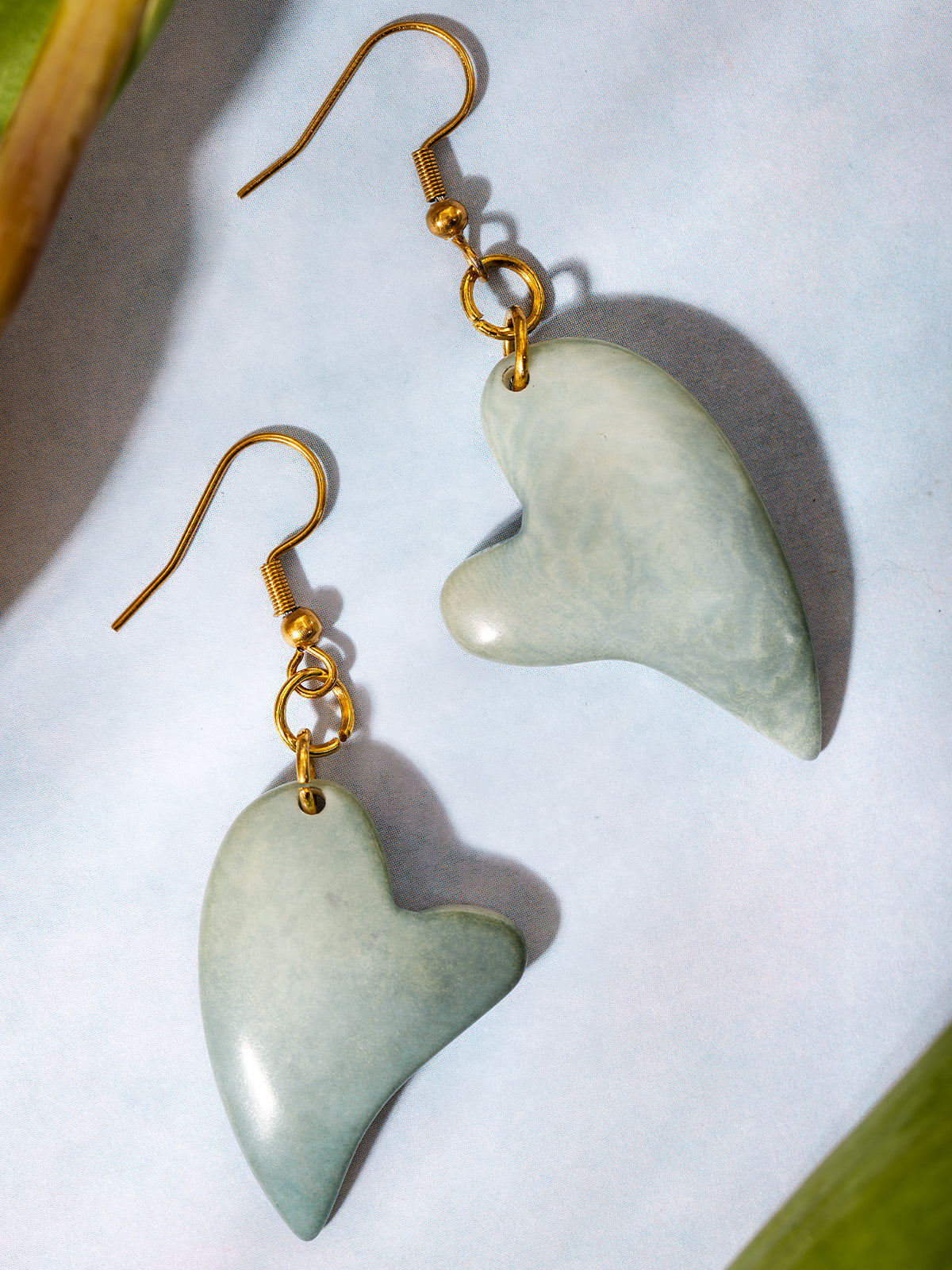 Handcrafted dangle earrings featuring asymmetrical heart-shaped green stone charms with gold hooks, displayed on a soft blue background.