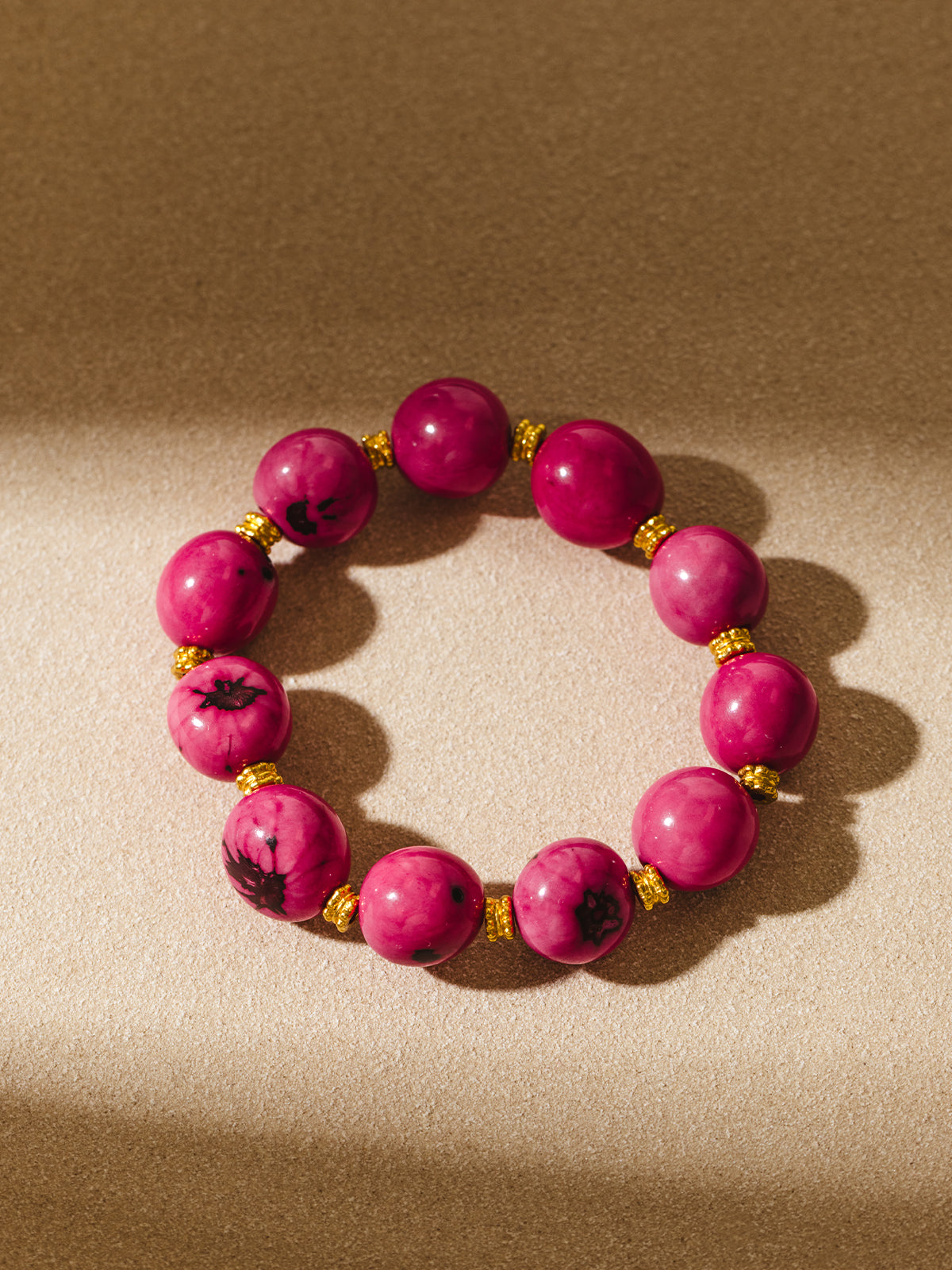 Flat lay of a bright pink beaded bracelet featuring large glossy round beads with dark marbling and gold spacer beads, displayed on a softly lit neutral surface.