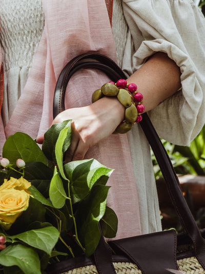 Close-up of a woman holding a woven market tote with yellow roses and greenery, wearing a cream dress and layered chunky bracelets in olive green and bright pink tones.