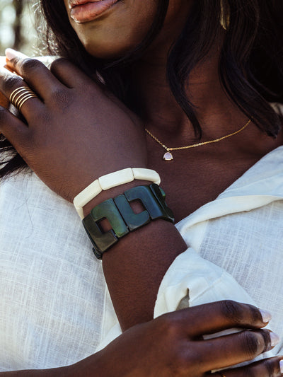 Elegant close-up of a woman wearing bold ivory and dark geometric bracelets, a gold necklace with a light pink pendant, and a multi-band gold ring, styled with a soft white linen blouse.
