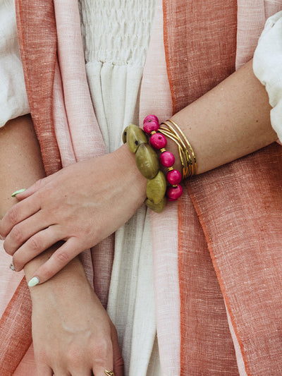 Close-up of a woman's hand and wrist adorned with gold bangles and chunky bracelets in olive green and bright pink, styled with a soft cream dress and a woven rust and blush scarf.
