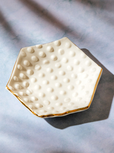 Hexagonal ceramic dish with raised dot texture and gold trim, displayed empty on a soft blue surface with gentle shadows.