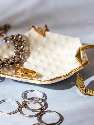 Close-up of a textured ceramic jewelry dish with gold trim, holding silver and gold rings, a gold cuff, and a chunky silver bracelet on a soft blue background.