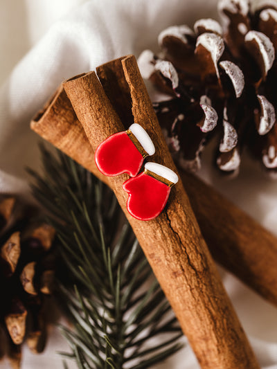 Red mitten stud earrings with white trim displayed on cinnamon sticks and pine cones – handmade holiday jewelry with cozy Christmas style.