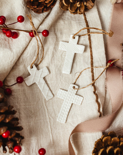 Set of three handcrafted white ceramic cross ornaments with textured patterns and jute hangers, styled on neutral linen with pinecones, red berries, and ribbon — artisan-made Christmas décor.