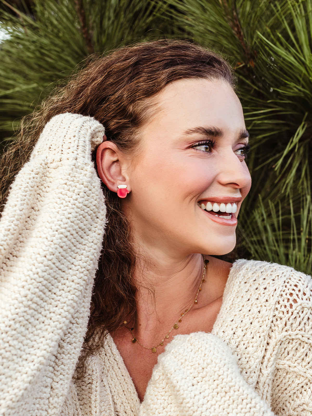 Smiling woman with curly hair wearing a cozy cream knit sweater and gold layered necklace, modeling handcrafted red and white stud earrings made by global artisans.