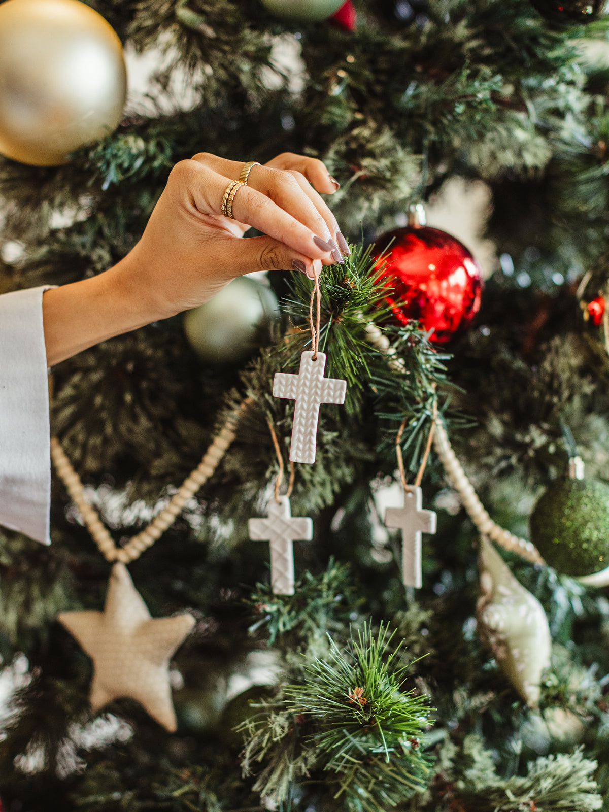Hand decorating a Christmas tree with white textured cross ornaments, surrounded by red, gold, and green baubles and wooden bead garland — ethically made holiday décor.
