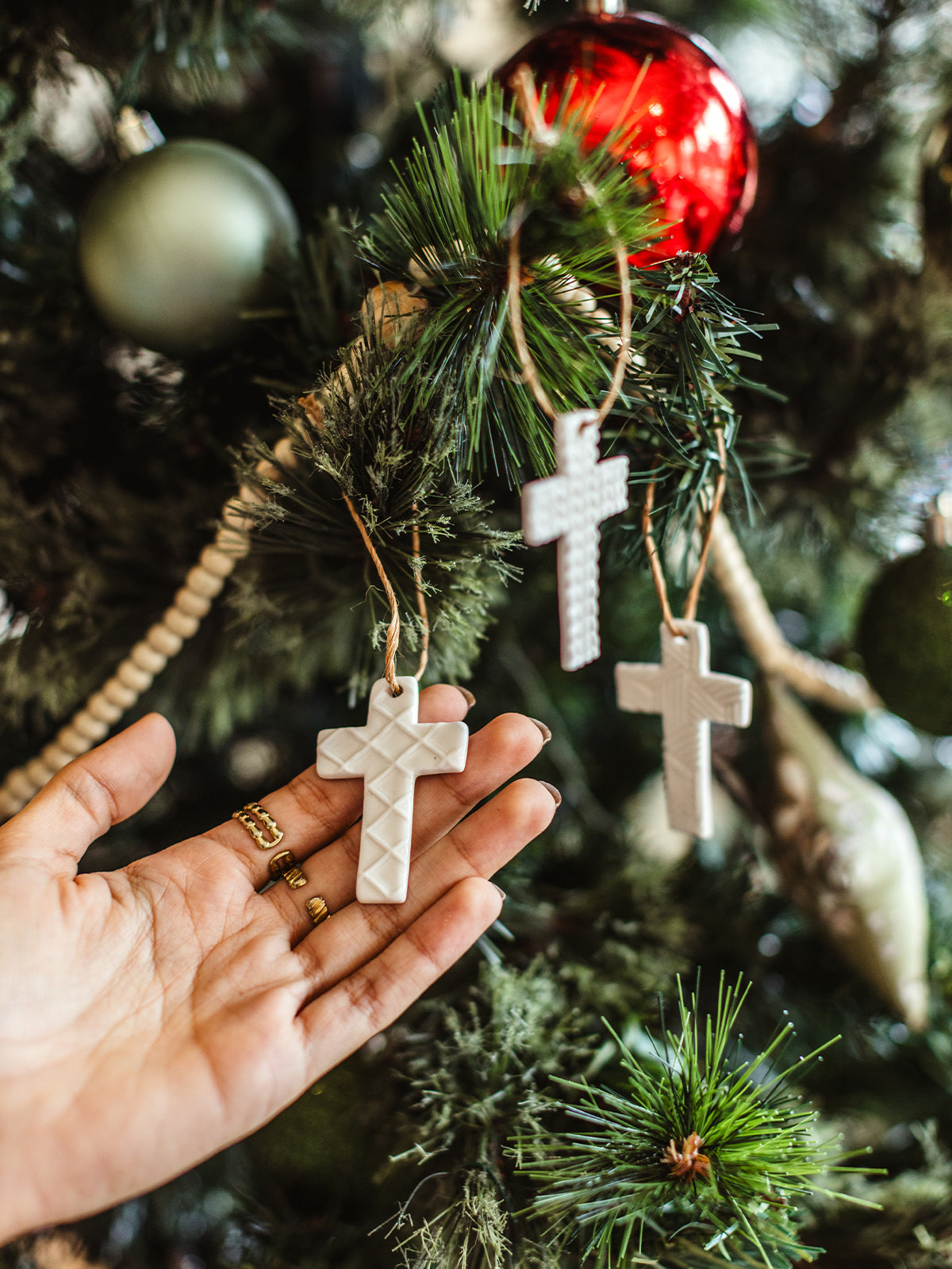 Close-up of hand holding a white ceramic cross ornament with a quilted texture on a Christmas tree, featuring red and green baubles — fair trade handmade holiday decoration.