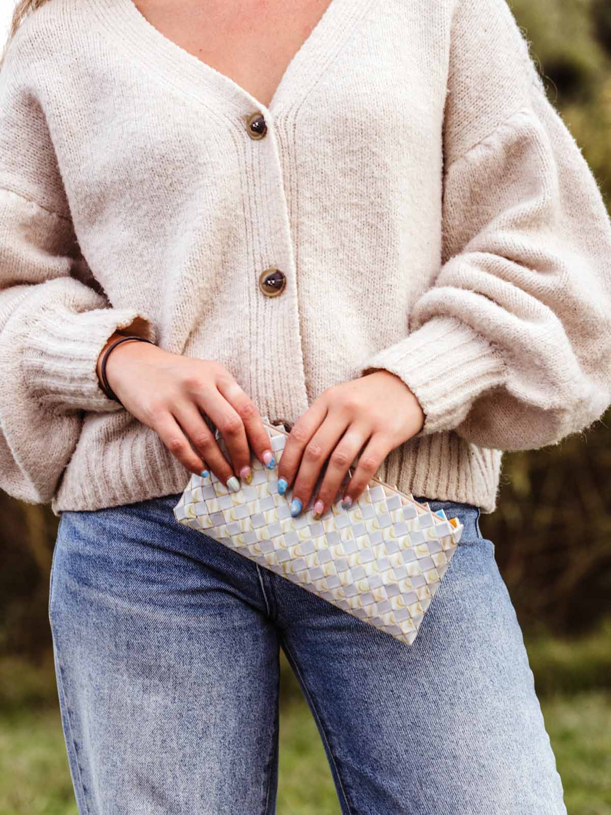 Close-up of a woman wearing a cozy beige knit cardigan and light-wash jeans, holding a handwoven patterned clutch in neutral tones.