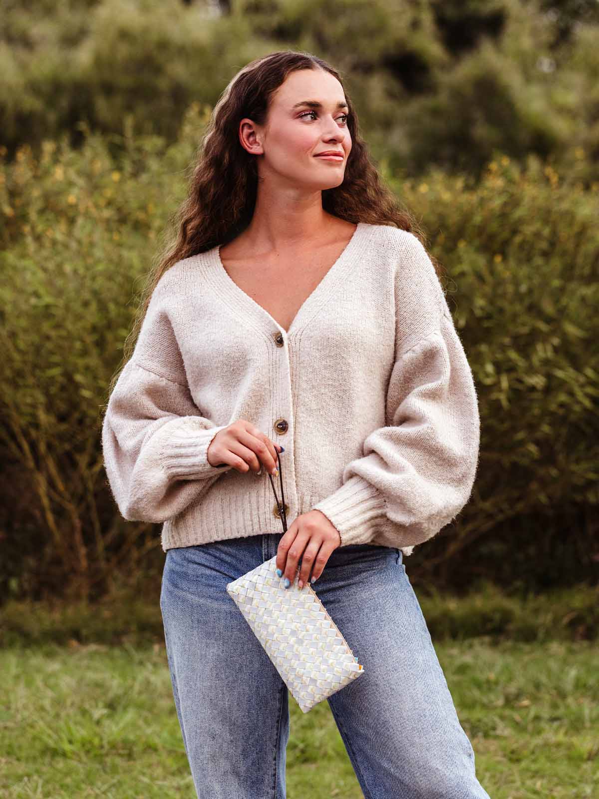 Woman standing outdoors wearing a beige knit cardigan and jeans, holding a woven clutch purse with a geometric pattern and looking to the side with a relaxed expression.