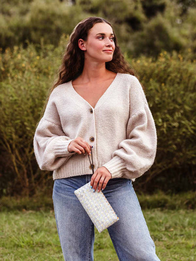 Woman standing outdoors wearing a beige knit cardigan and jeans, holding a woven clutch purse with a geometric pattern and looking to the side with a relaxed expression.