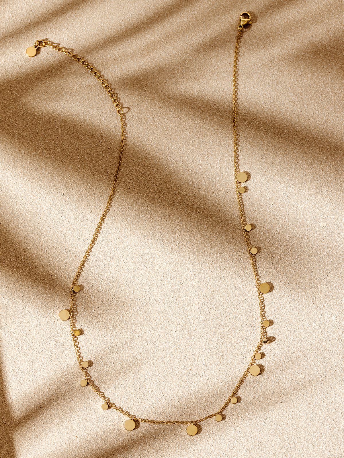 Minimalist gold charm necklace with scattered disc accents, arranged on a textured beige backdrop with warm natural shadows.
