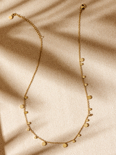 Minimalist gold charm necklace with scattered disc accents, arranged on a textured beige backdrop with warm natural shadows.