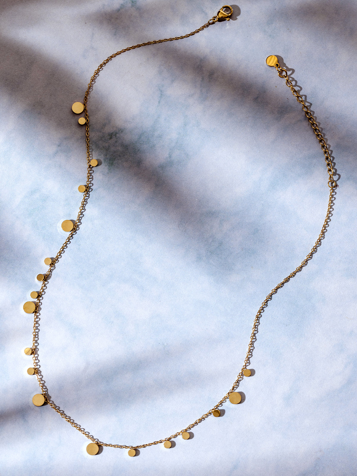 Gold disc charm necklace laid out in an elegant curve on a dreamy blue-toned surface, showcasing the shimmering detail of the varying charm sizes.