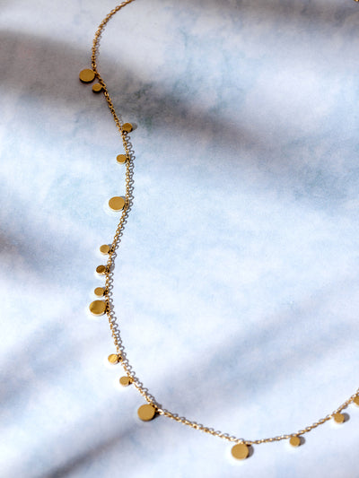 Delicate gold chain necklace with small and large flat disc charms, displayed in soft lighting on a cloud-like blue and white background.
